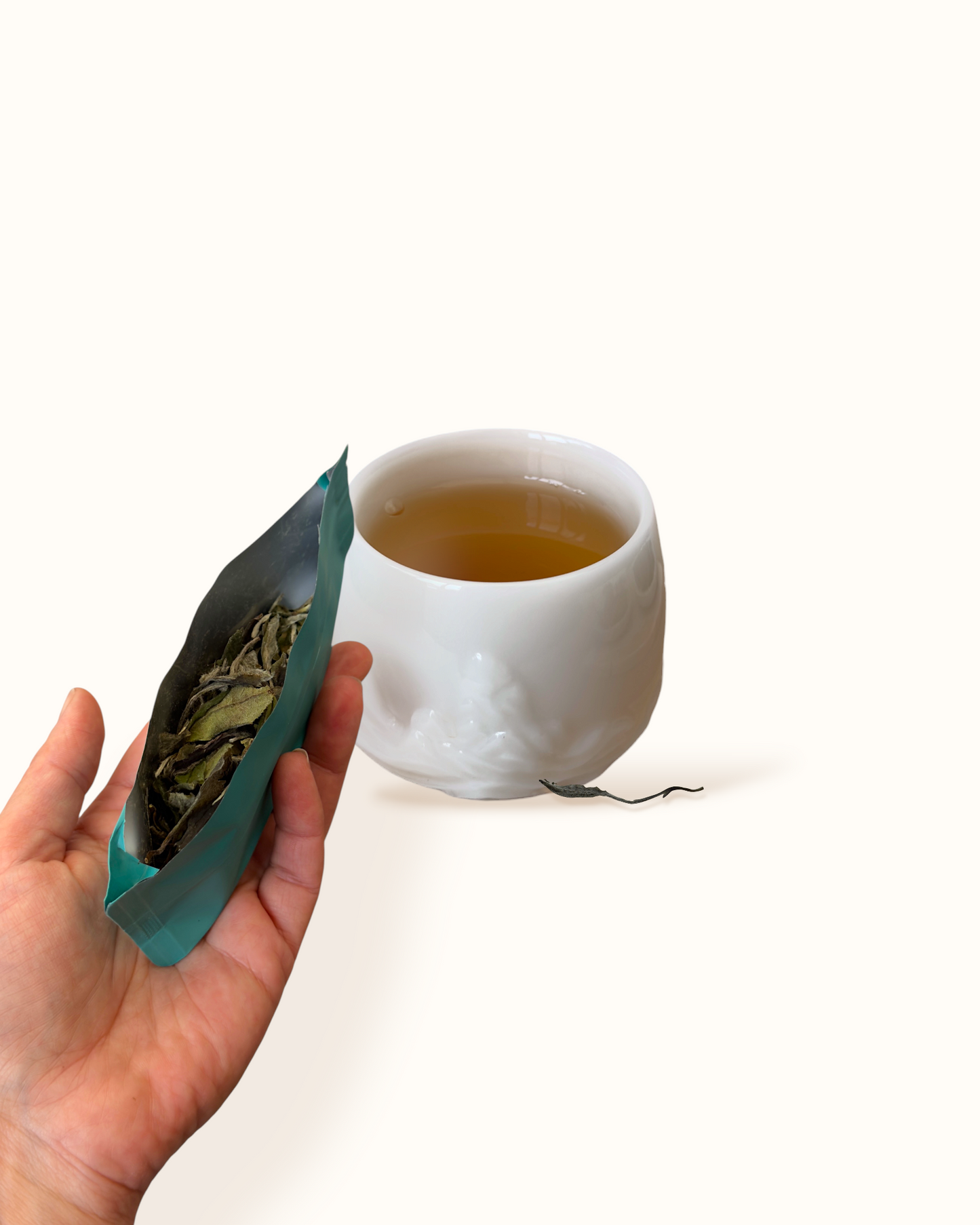 Close-up of White Peony white tea leaves with an open teal sachet in hand beside amber-golden tea liquor in a white ceramic tea cup
