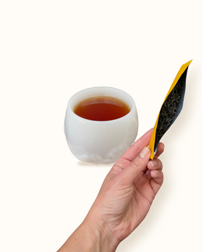 Close-up of Lapsang Souchong black tea leaves with an open orange sachet in hand beside dark amber tea liquor in a white ceramic tea cup
